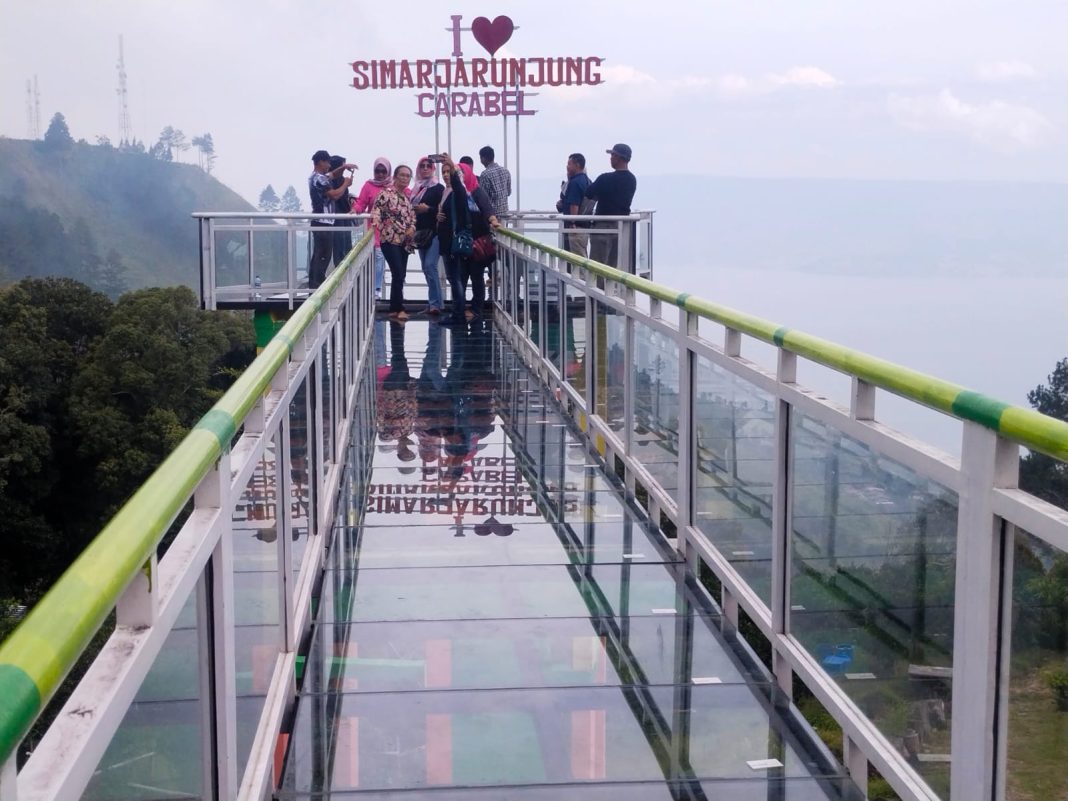 Para wisatawan sedang asyik berswafoto di jembatan kaca Carabel Simarjarunjung Kabupaten Simalungun dengan latar belakang pemandangan alam Danau Toba, Minggu (6/8/2023).