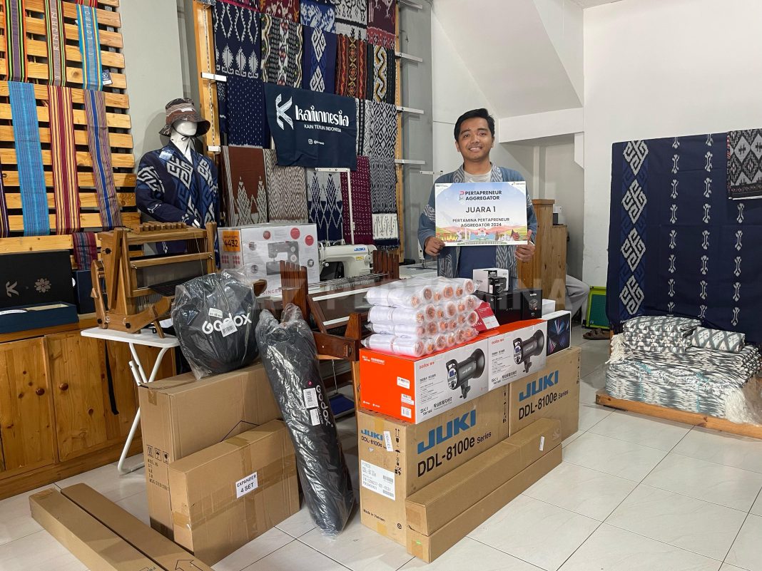 Salah satu pemenang Pertapreneur Aggregator 2024 berpose dengan berbagai peralatan teknologi hibah dari PT Pertamina (Persero) di tengah koleksi kain tenun Indonesia. (Dok/Pertamina)